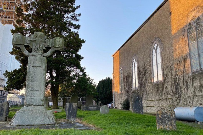 A cemetery with a large stone cross, a church with arched windows, and a tree in the background. 2