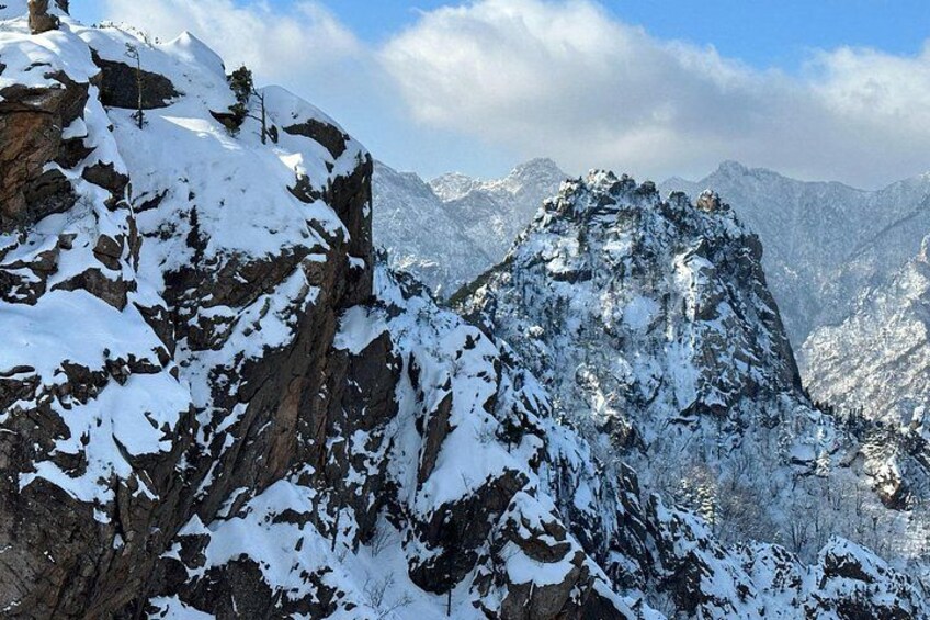 A snow-covered mountain peak with rocky terrain and distant mountains under a partly cloudy sky. 4