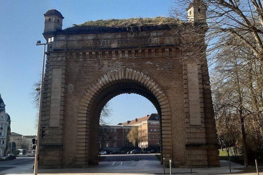 A large brick archway with inscriptions, surrounded by a paved area and leafless trees. 2