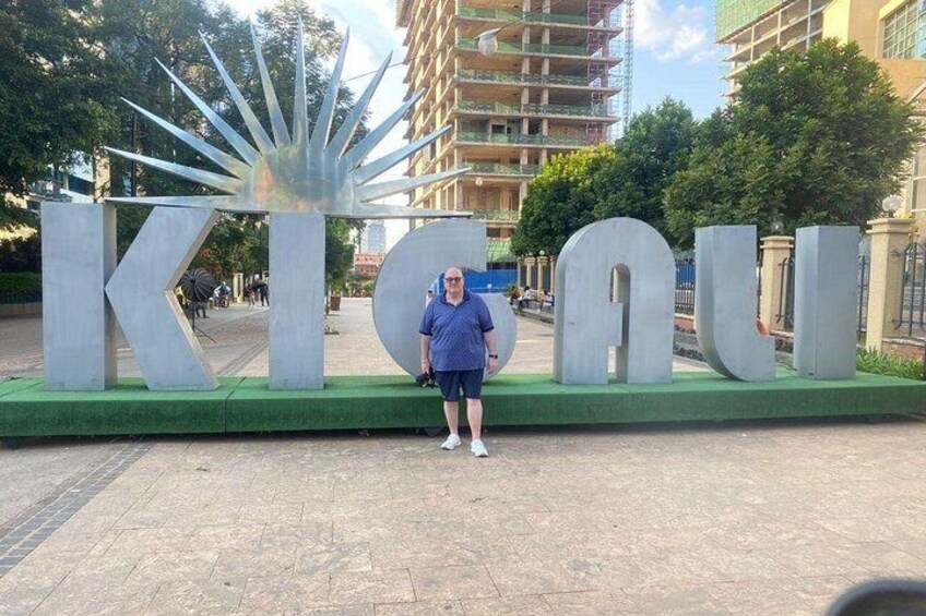 A person standing in front of large metallic letters spelling 'KICOU' with a building in the background. 1