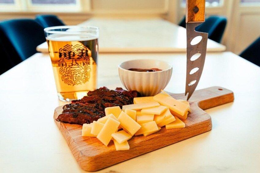 A wooden cutting board with cheese and meat, a glass of beer, and a knife on a table. 2