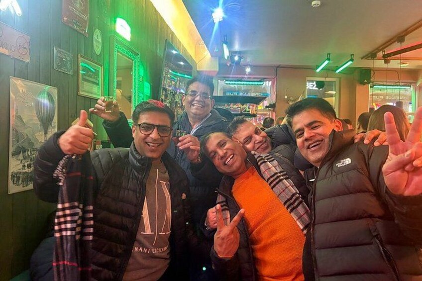 A group of people in a warmly lit indoor setting with green ambient lighting, some holding drinks, and a visible sign with the word 'BEZ'. 5