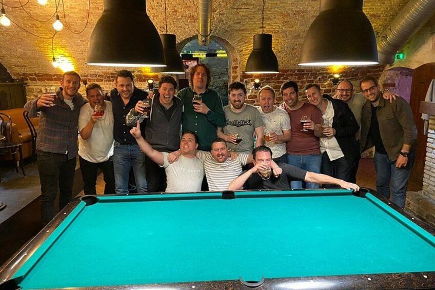 A group of people around a pool table in a rustic indoor setting with exposed brick walls and hanging pendant lights. 1