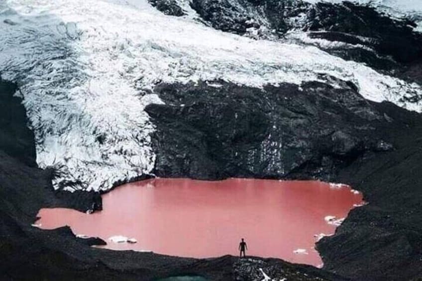 Une personne se tient au bord d’un lac de cratère rouge, entouré de neige et d’un paysage volcanique. 3