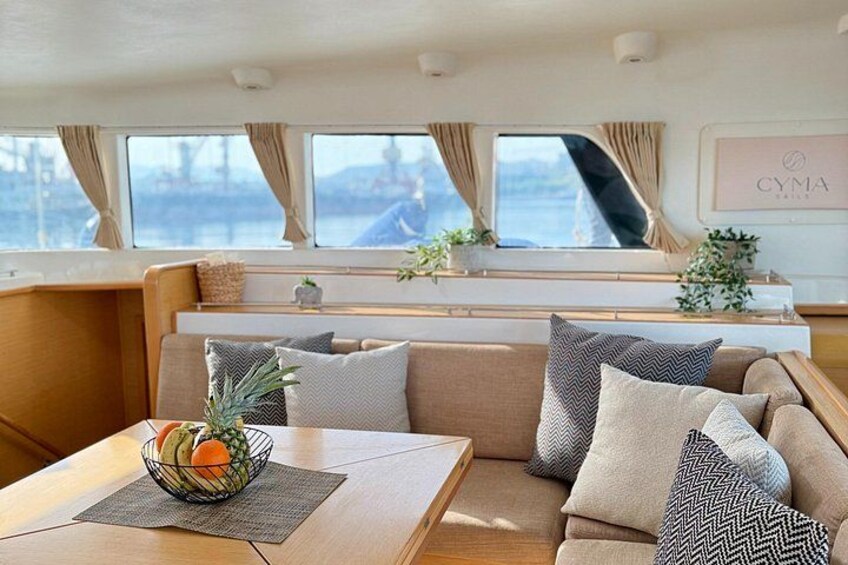 A cozy cabin interior with a sofa, cushions, a table with a fruit bowl, and a window overlooking a body of water. 3
