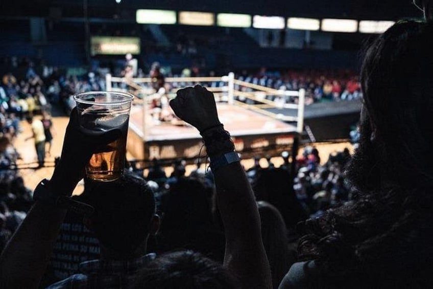 Una persona sosteniendo una cerveza en un ambiente interior tenuemente iluminado, probablemente una arena deportiva con un ring de boxeo visible al fondo. 4