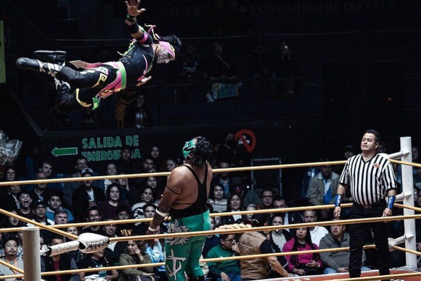 A wrestling match with a referee and two wrestlers in a ring, surrounded by an audience. 1