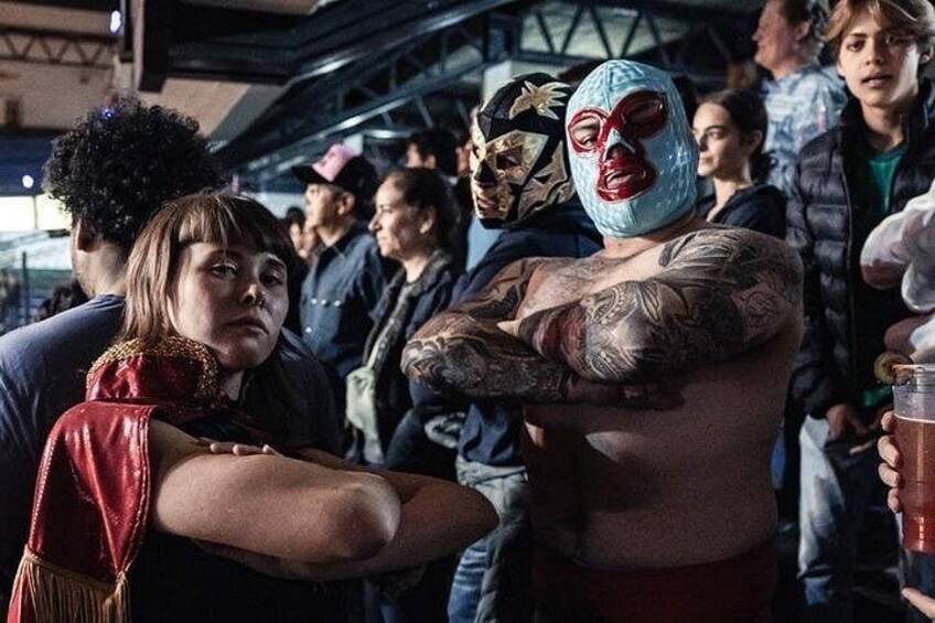 Una persona con tatuajes y una máscara, de brazos cruzados, en un evento concurrido. 2