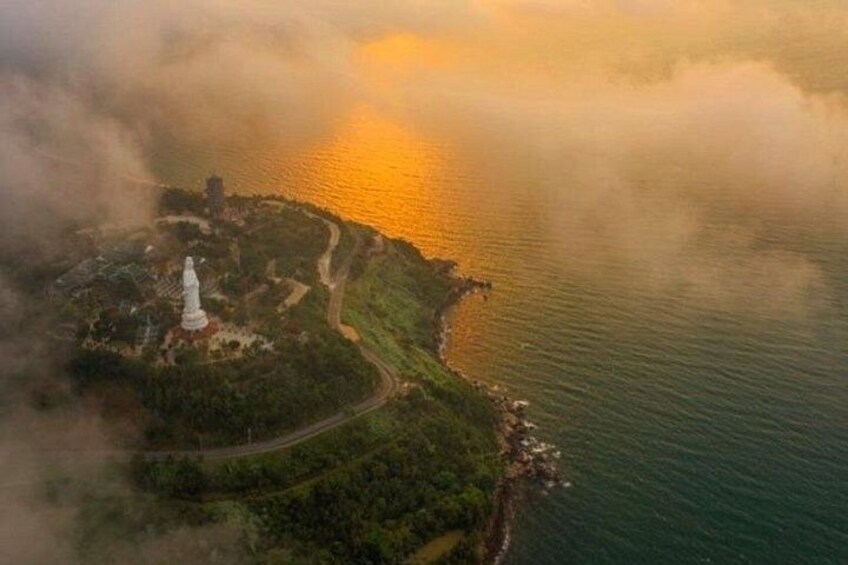 An aerial view of a coastal area with a prominent white statue, a winding road, and a body of water reflecting sunlight. 4