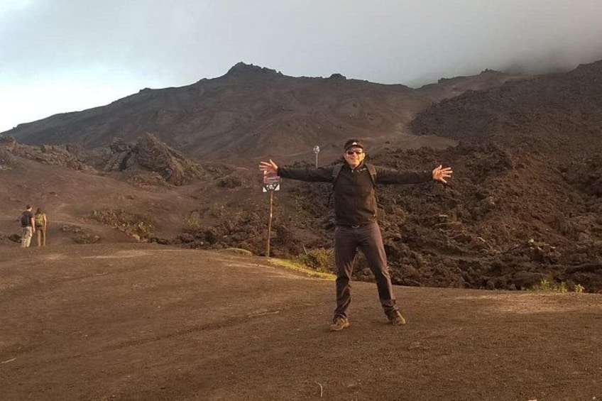 A person standing on a dirt road with arms outstretched, a mountainous landscape in the background, and another person walking in the distance. 2