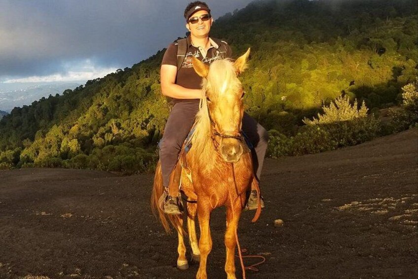 Una persona montando a caballo por un sendero de tierra con un paisaje montañoso cubierto de bosque al fondo. 3