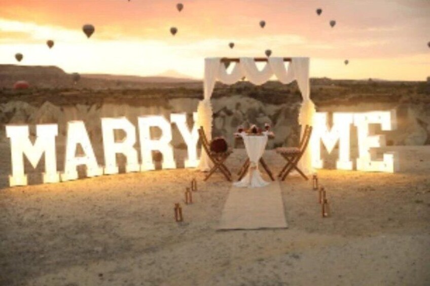 A desert wedding setup with a 'MARRY ME' sign, a white canopy, and a table with guests. 2