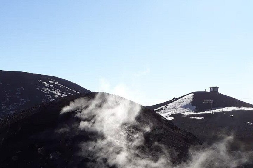 Paisaje volcánico con vapor ascendiendo del suelo, una montaña nevada al fondo y una pequeña estructura en la cima. 1