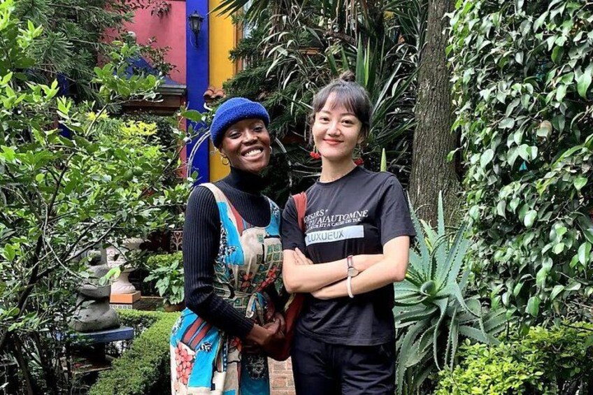 Two people standing in a garden with dense foliage and a colorful building in the background. 3