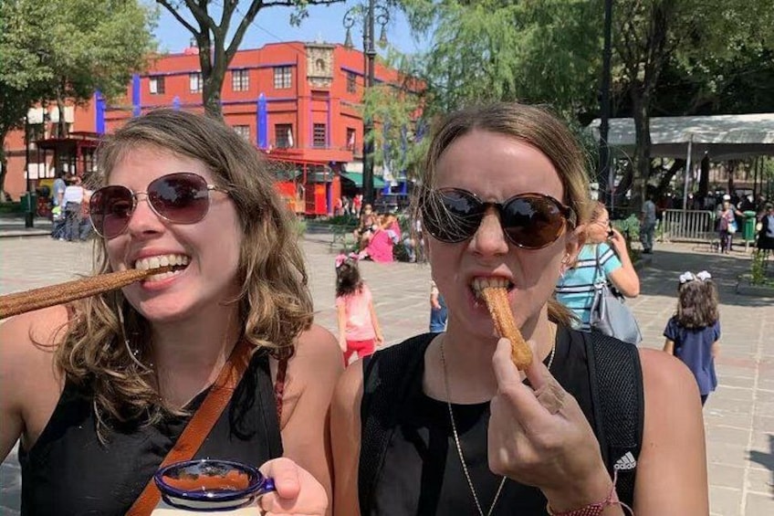 Two people eating churros in an outdoor setting with buildings and trees in the background. 1
