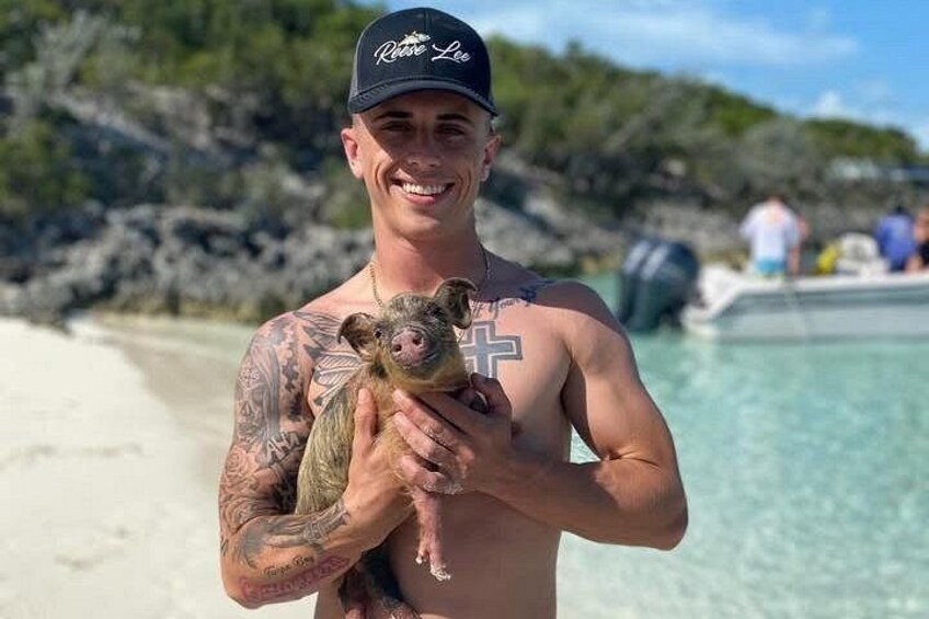 A person holding a piglet on a beach with clear water and rocky terrain in the background. 4
