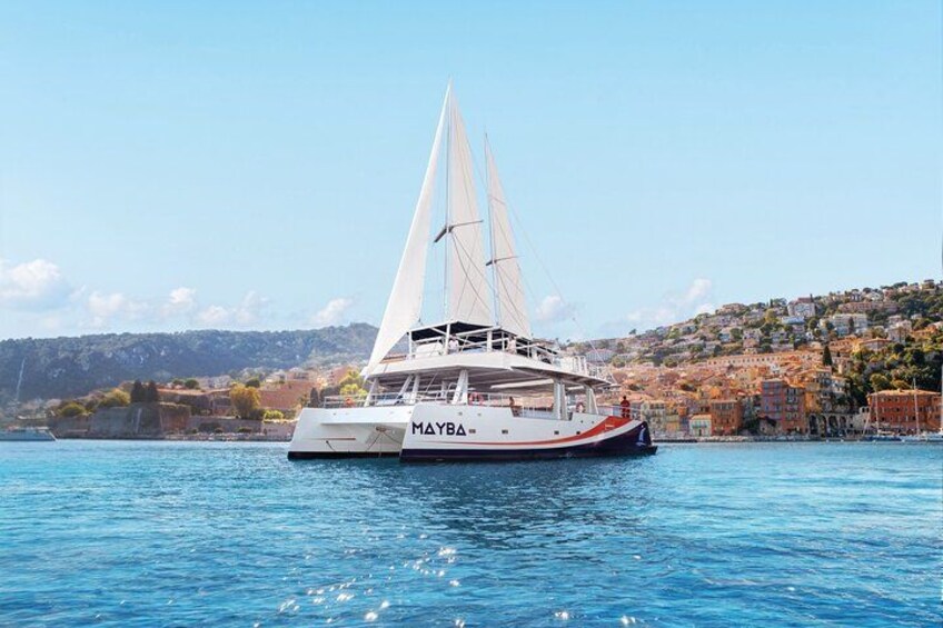 A white catamaran named MAYBA sailing on calm waters with a coastal town in the background. 2