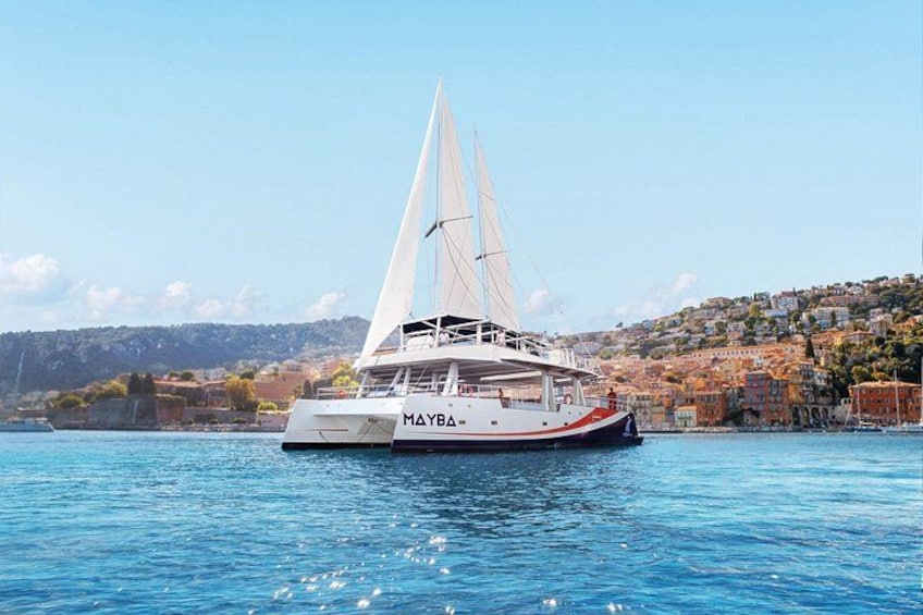 A white catamaran named MAYBA sailing on calm blue waters with a coastal town in the background. 3
