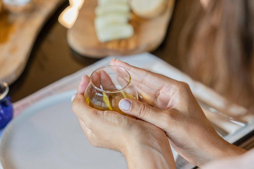 Hands holding a small glass with a yellow liquid, possibly tea, on a table with a white plate and a wooden tray with sliced bread in the background. 2