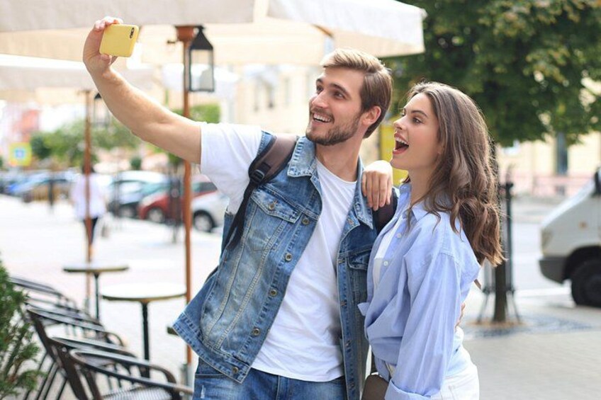 A person taking a selfie with another person standing beside them, a street with parked cars, and outdoor seating. 2