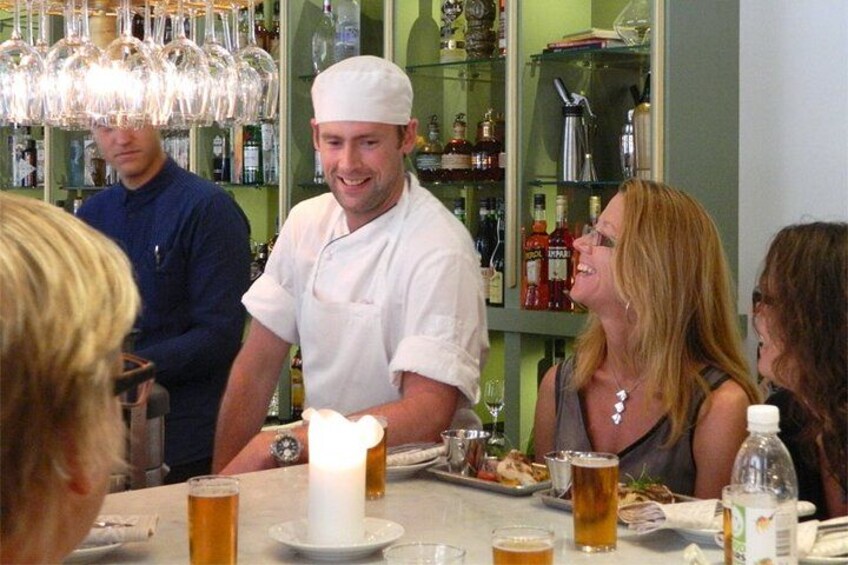 Ein Restaurant mit einem Kellner, der Speisen serviert, einer Kerze auf dem Tisch und verschiedenen Flaschen und Gläsern. 1