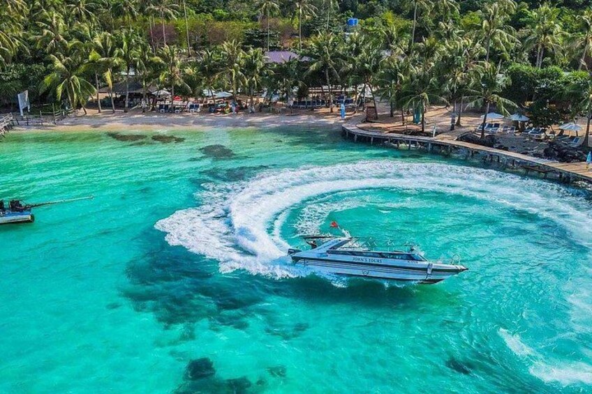 Een boot die een kielzog achterlaat in turquoise water, omringd door palmbomen en een strand met een pier. 1