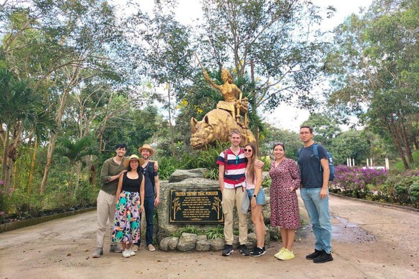 Eine Gruppe von Personen steht vor einer Statue mit einer Gedenktafel, umgeben von Grünpflanzen. 2
