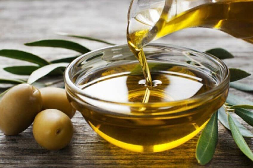 Olive oil being poured into a glass bowl with olives and olive leaves in the background. 1