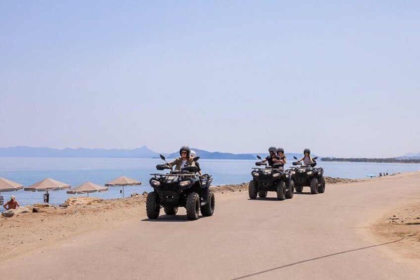 Three people riding ATVs on a beach road with umbrellas and mountains in the background. 4