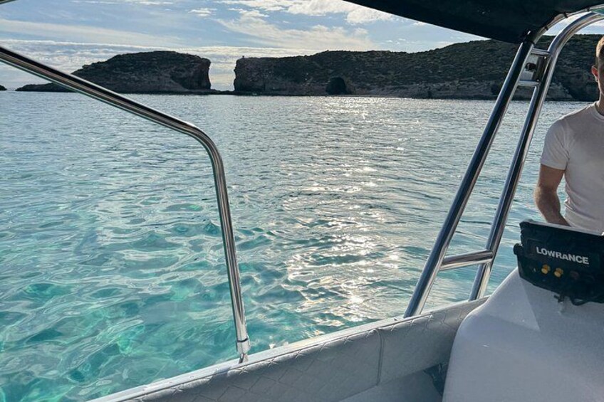 A boat with a person wearing a white shirt and a black bag labeled 'LOWRANCE' on the deck, with clear turquoise water and rocky formations in the background. 3