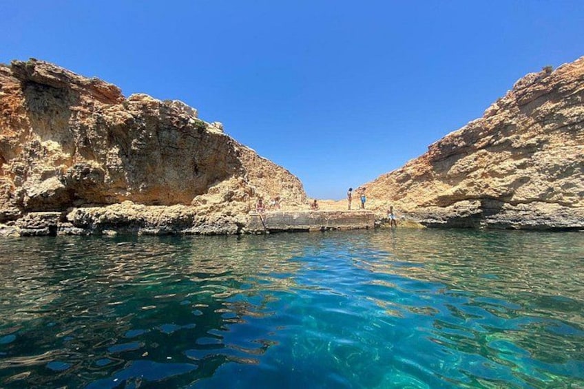 A coastal landscape with clear blue waters, rocky cliffs, and a few people near the shore. 4