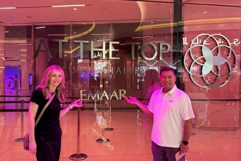 Two people standing in front of a glass storefront with the words 'AT THE TOP' and 'EMAAR' visible. 1