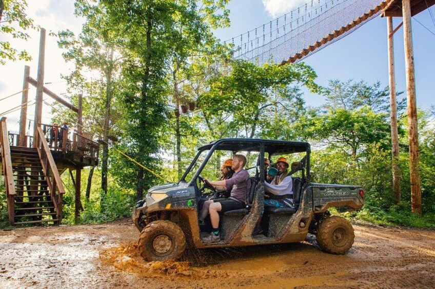 A four-wheeler vehicle on a muddy path with a wooden bridge and trees in the background. 4