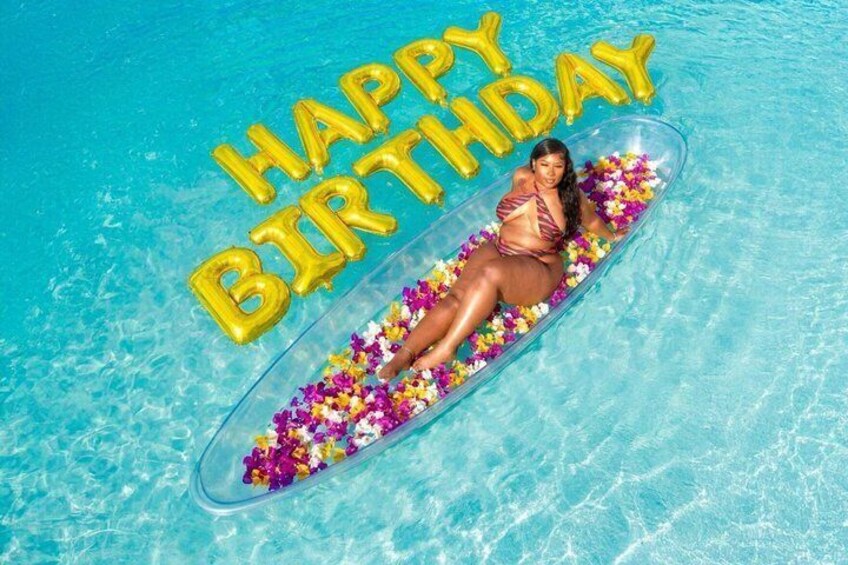 A person floating on a clear inflatable raft in a pool with 'HAPPY BIRTHDAY' balloons. 1