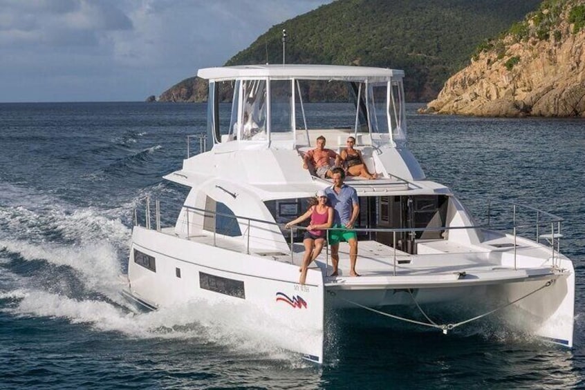 Eine weiße Yacht mit Sonnensegel, Personen auf dem Deck und eine felsige Küstenlinie im Hintergrund. 1
