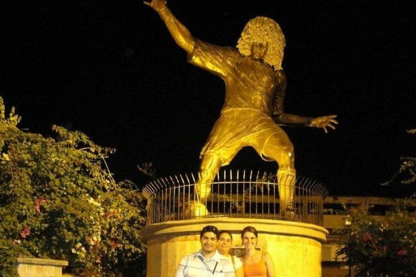 En gyllene staty av en figur med utsträckta armar, omgiven av ett staket och grönska. 3