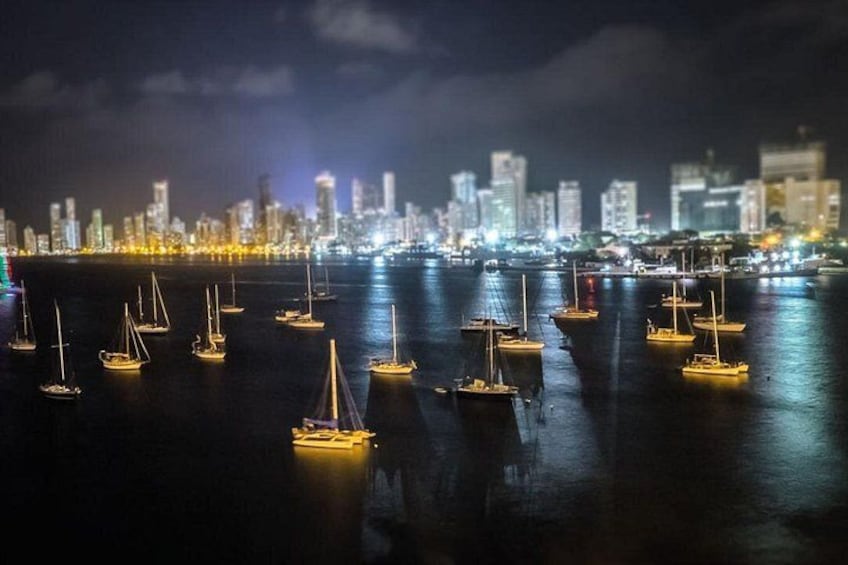 Una marina nocturna con veleros anclados en aguas tranquilas, un horizonte urbano al fondo y un puente que cruza el agua. 5