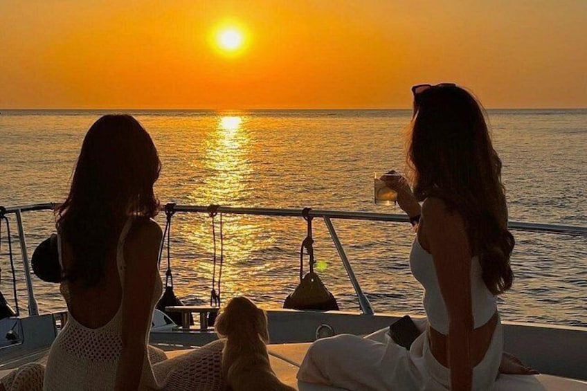 Twee vrouwen zitten in een boot, waarvan er één een drankje vasthoudt, met een zonsondergang boven de oceaan. 4