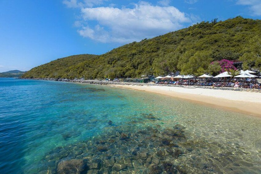 A beach with clear turquoise water, a sandy shore, and a forested hill in the background. 5