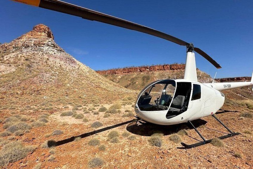 A white helicopter with 'JA-ATN' on its tail rotor, parked on a rocky, arid landscape with sparse vegetation and a prominent red rock formation in the background. 3