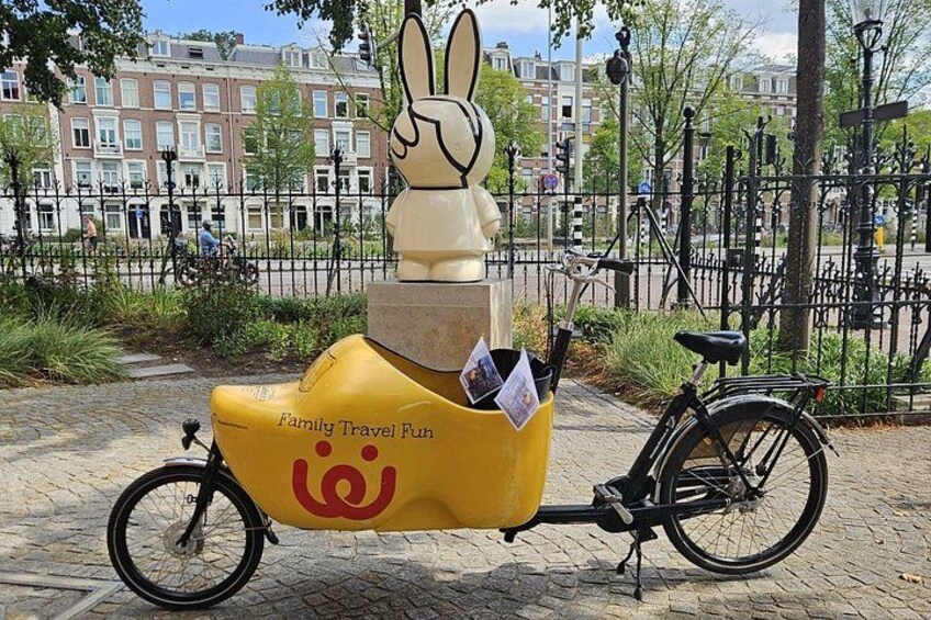 A yellow bicycle with a rabbit sculpture on the seat, parked on a cobblestone path. 1