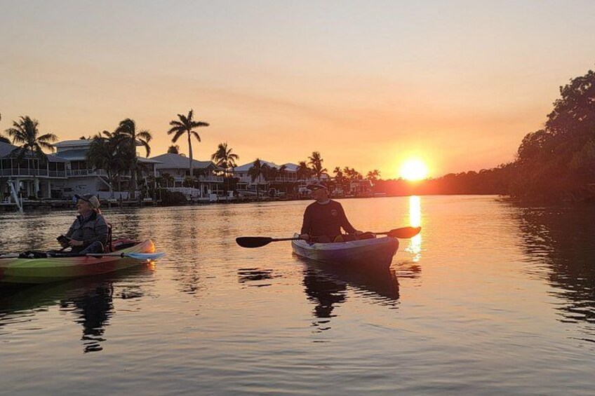 Dos kayakistas en un río tranquilo al atardecer, con palmeras y casas al fondo. 3