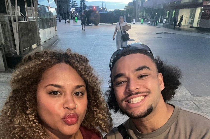 Two people taking a selfie on a city street with a construction site and a police vehicle in the background. 2
