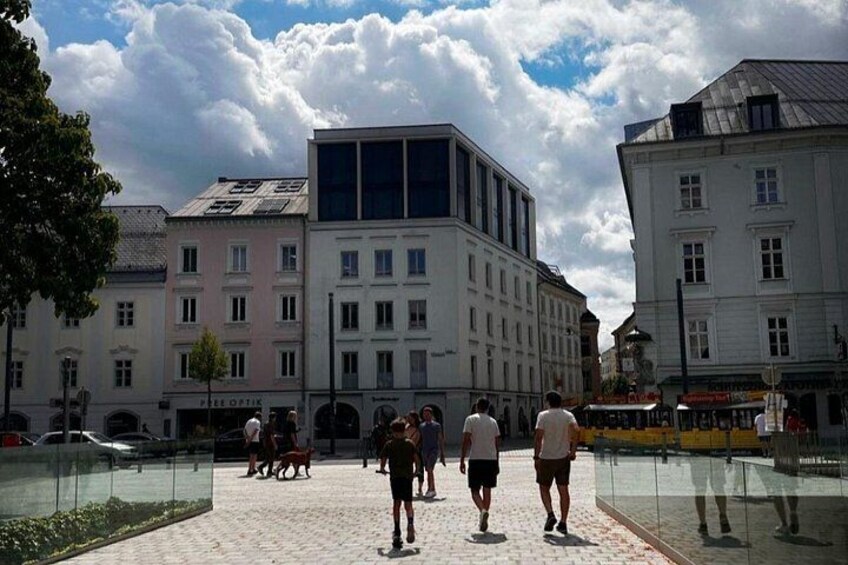 A pedestrian area with buildings, a glass enclosure, and people walking. 1