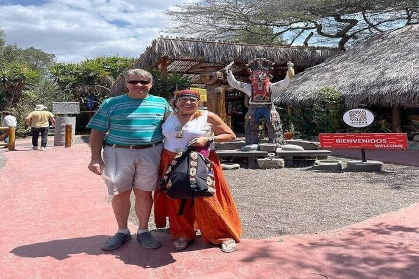 A couple standing in front of a welcome sign, a thatched-roof structure, and a statue. 5