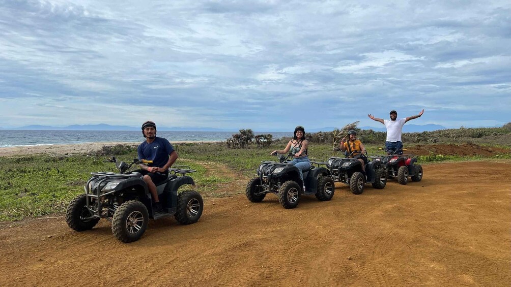Four people on ATVs on a dirt path near a beach with the ocean in the background. 4