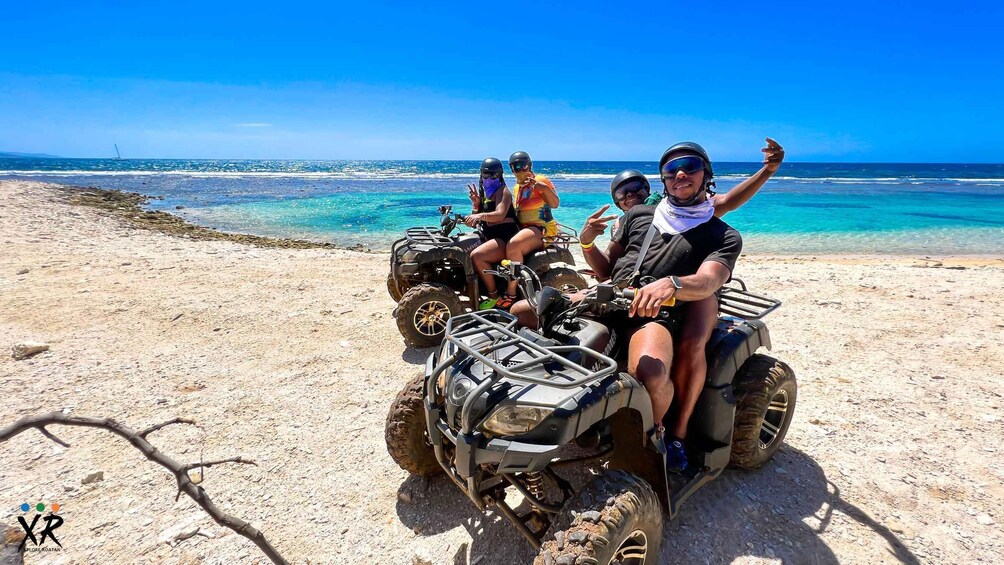 A group of people on ATVs on a sandy beach with clear blue water and a boat in the distance. 1