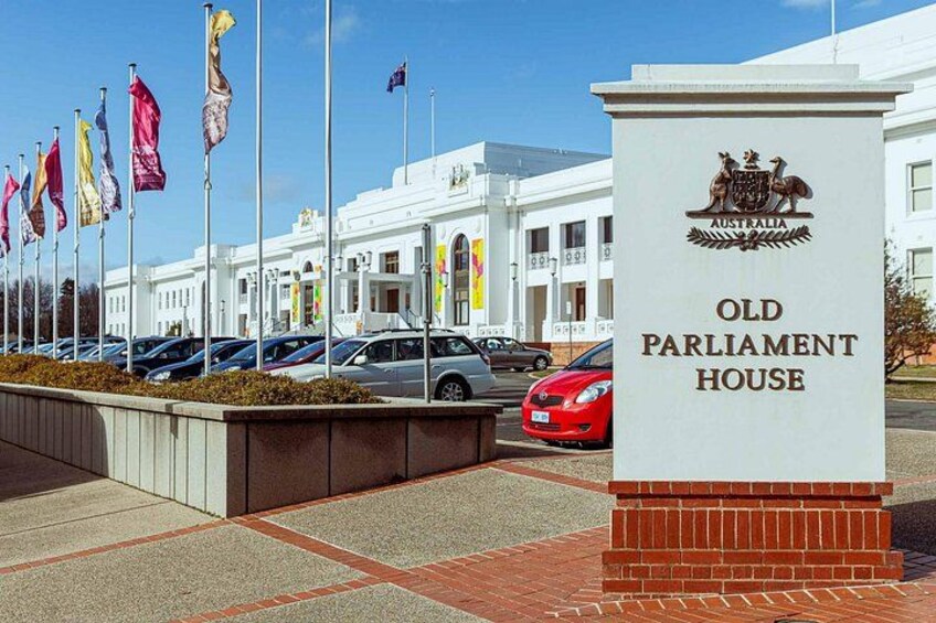 The Old Parliament House with flags, a parking lot, and a clear blue sky. 1