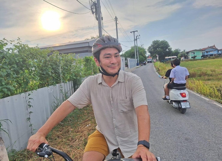 A person on a bicycle and another on a scooter on a road with houses and greenery in the background. 1