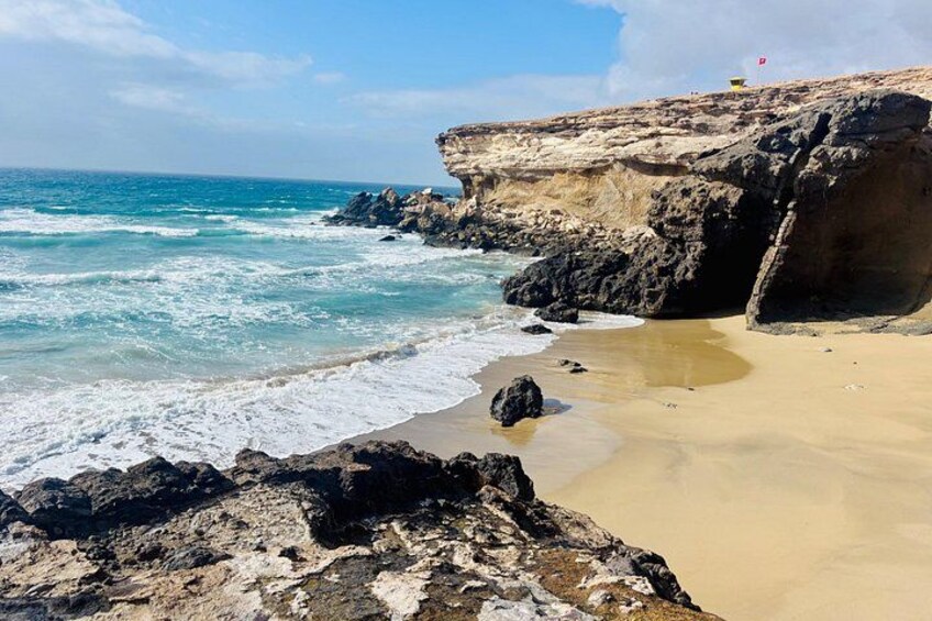 A rocky coastline with waves crashing onto the shore, a sandy beach, and a flag on a cliff. 2